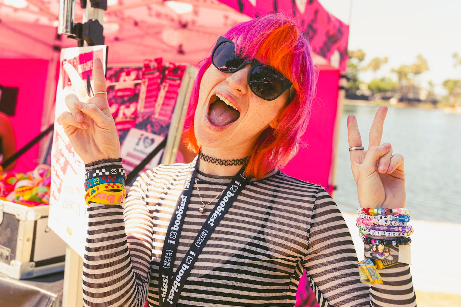 Person with pink hair and sunglasses on, standing in front of the i love boobies! booth and wearing a bunch of bracelets from the brand.
